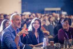 Photo of Josh Clark speaking in a microphone at a table among a large listening audience